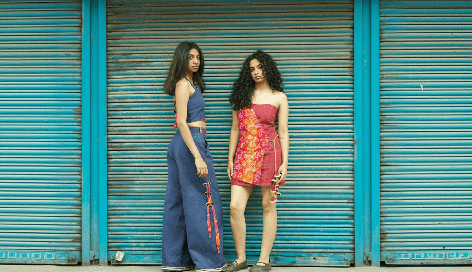 Two models in blue and red outfits in front of turquoise shutters